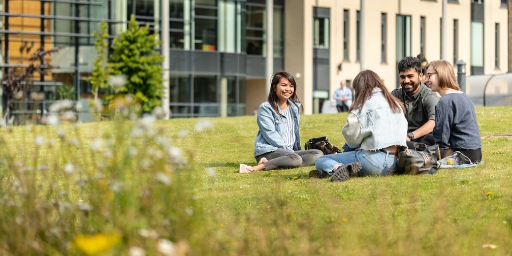 A group of students sat on grass on campus on a sunny day with buildings in the background.
