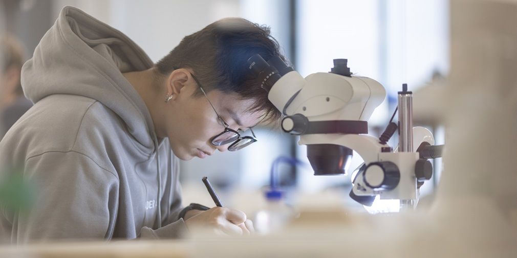 A student sat in a lab, writing. A microscope is in the foreground