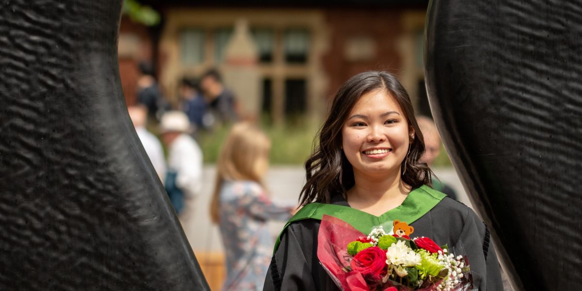 A student in graduation robes, holding a bunch of flowers. She is stoofd behind the 'Sign For Art sculpture on campus, and part of the black columns of the sculpture are in the foreground.