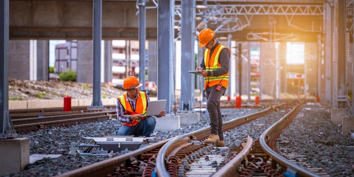 Two engineers in hi-vis and safety gear on a railway line. One stands with a clipboard and the other kneels, pointing at a notepad in their hand.