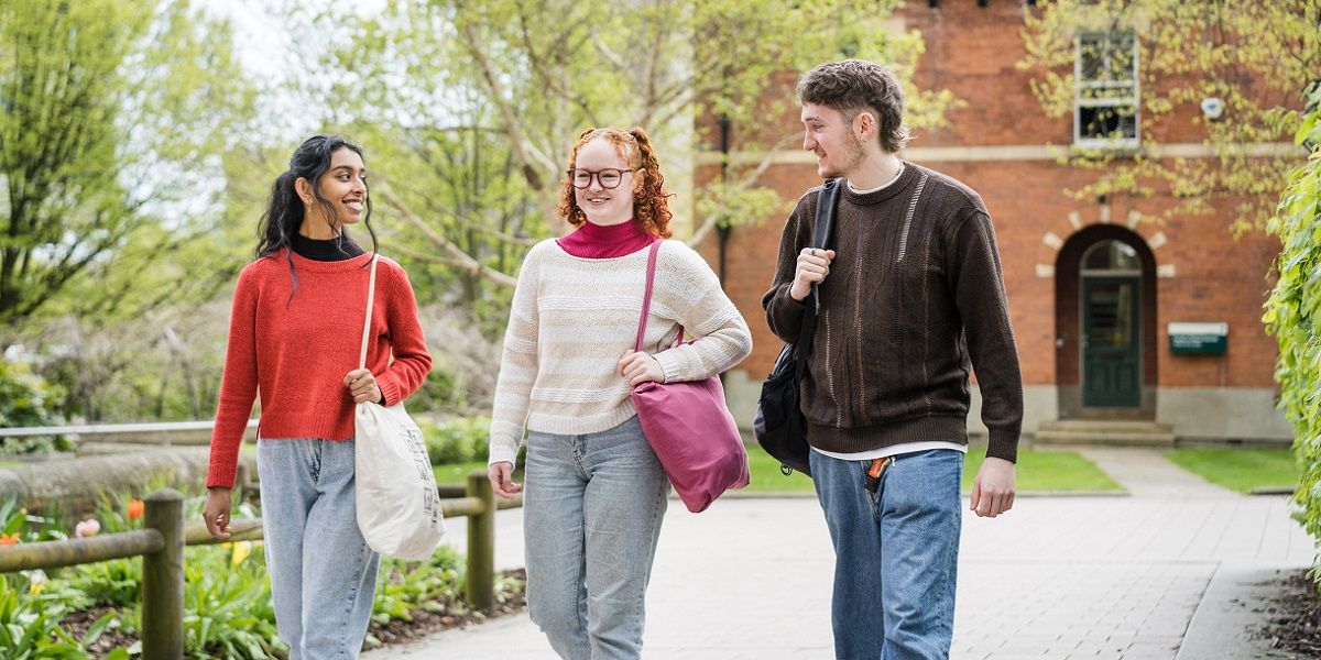 Three students walking on campus, smiling and chatting, with flowers, trees and a red brick building in the background.