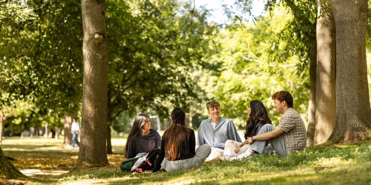 A group of students sit on the ground in a park on a sunny day, chatting.