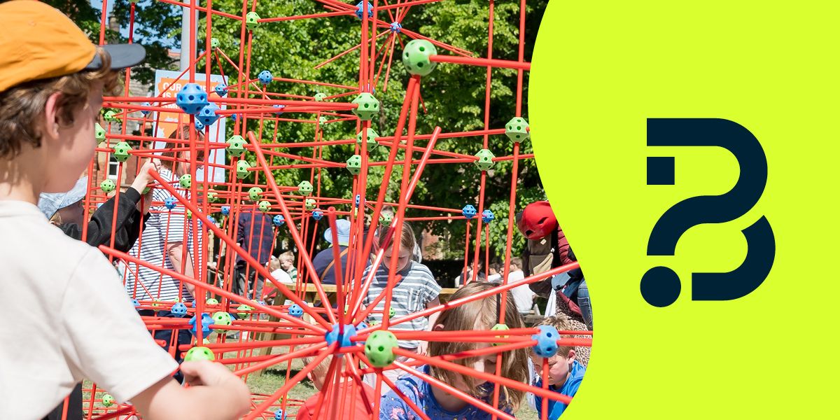 Children interact with a large red geometric climbing structure at an outdoor event, with a green panel on the right, featuring the Be Curious logo.