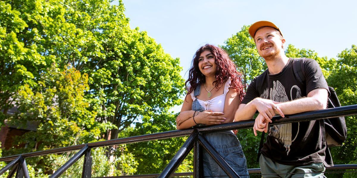 A group of students chatting and smiling, sat on a bench with trees and old buildings in the background.
