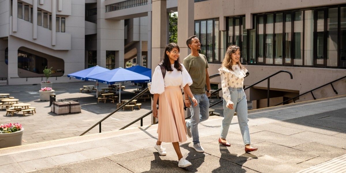 Landscape angled view of three students walking at the top of some stone steps outside the Edward Boyle Building. They are all dressed in summer clothing and the day is bright and sunny.