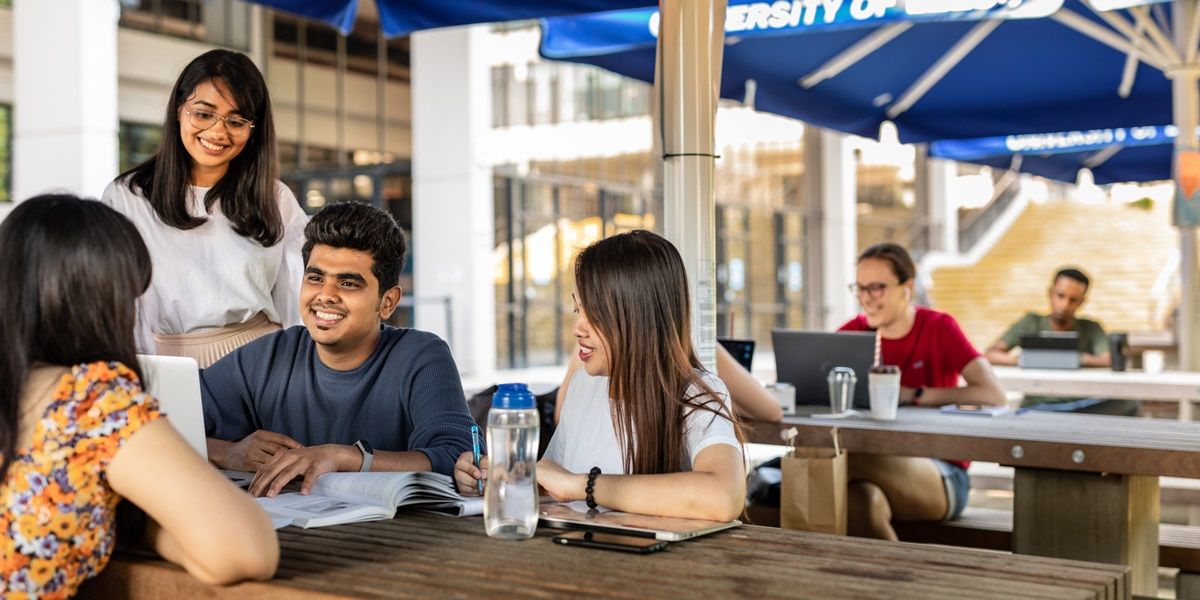 A group of students gathered around a picnic table on a sunny day on campus, chatting.