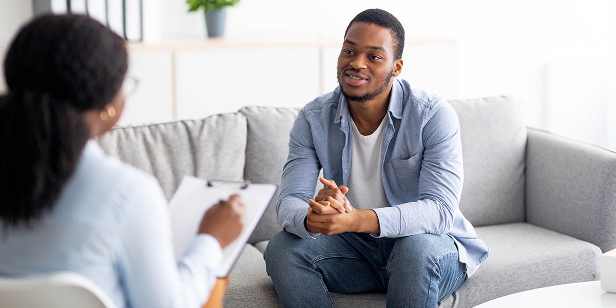 A man sat on a sofa talking to a clinician who is making notes on a clip board.