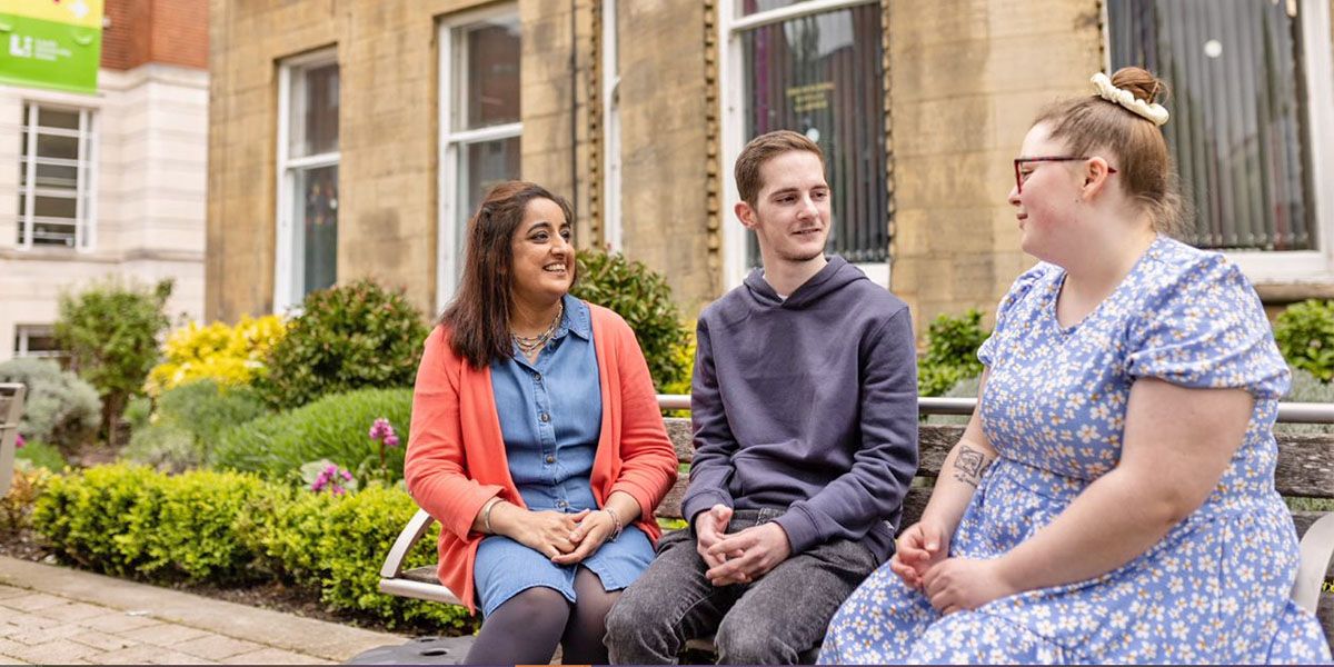 Three mature students sat smiling while seated on a bench. Flowers on campus are visible beyond.