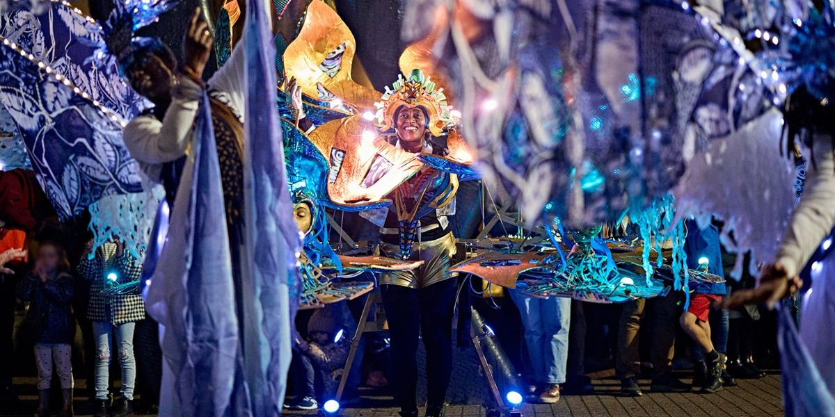 A smiling person in a flamboyant carnival costume which is gold and appears to glow against other people and costumes surrounding them.