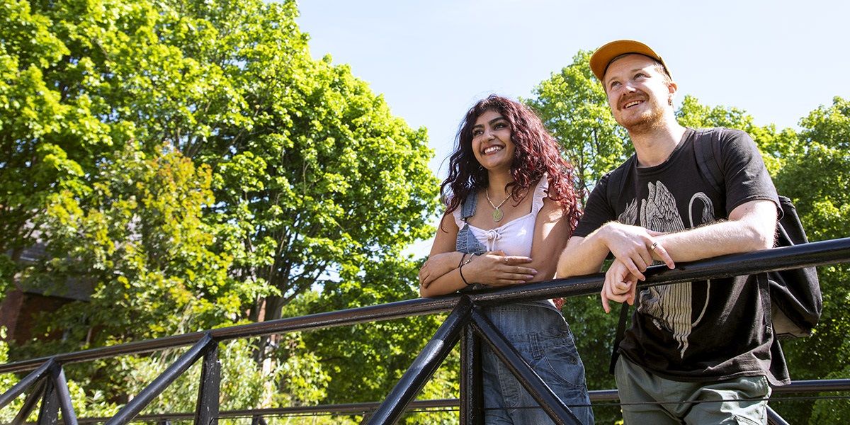 Two smiling students stood on a balcony outside on campus with trees in the background.