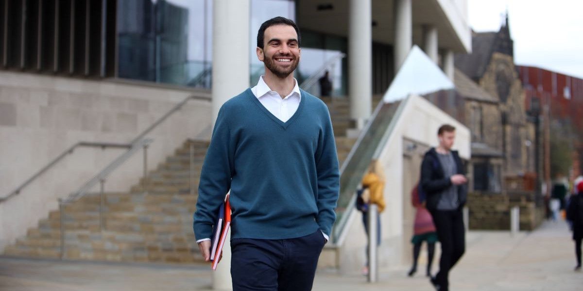A person walks and smiles while carrying two notebooks in their right hand. There are the steps up to an entrance of a modern building behind them and other students walk about in the background.
