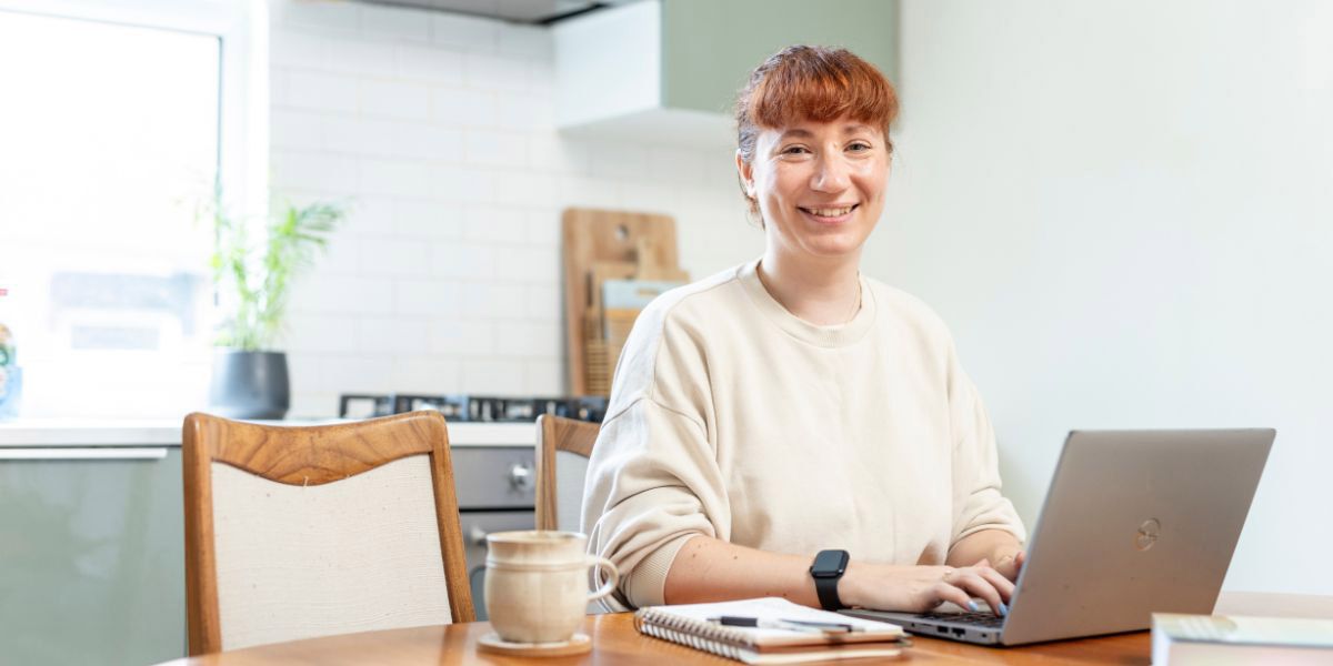 Masters student sat at a kitchen table using a laptop to study. They are smiling.