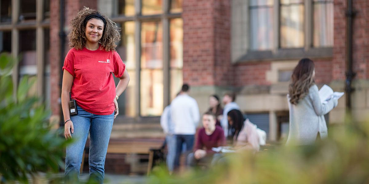 A student representative stood on campus smiling. Several visiting students are exploring the red-brick building and grounds beyond.