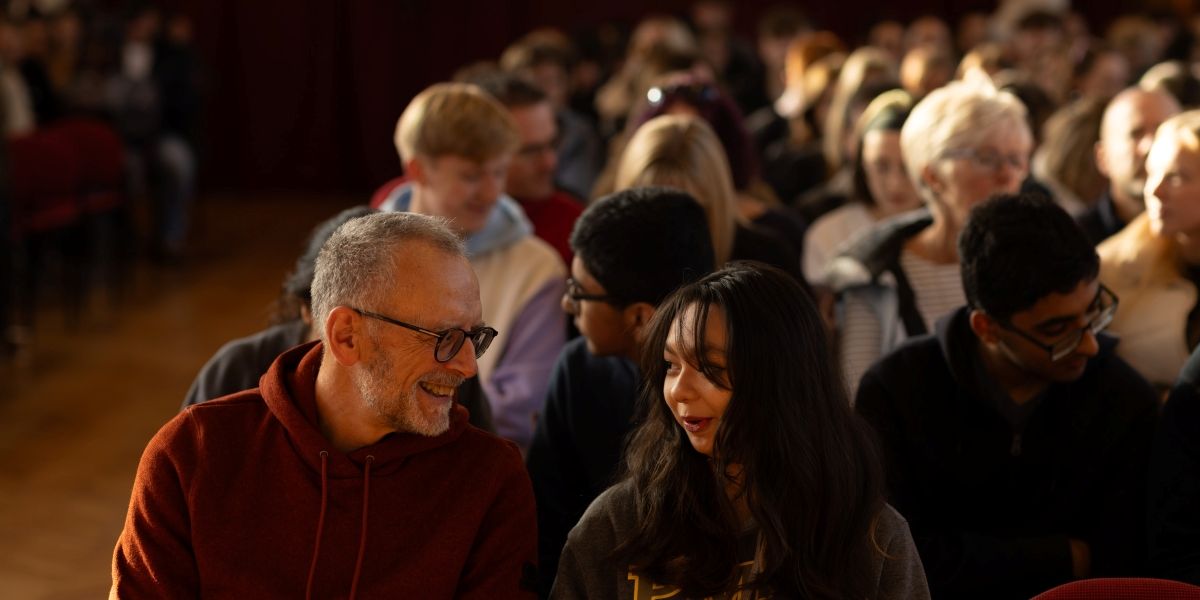 A parent and young person sit next to each other chatting. Behind them are rows of people as they wait for a talk in the Great Hall.