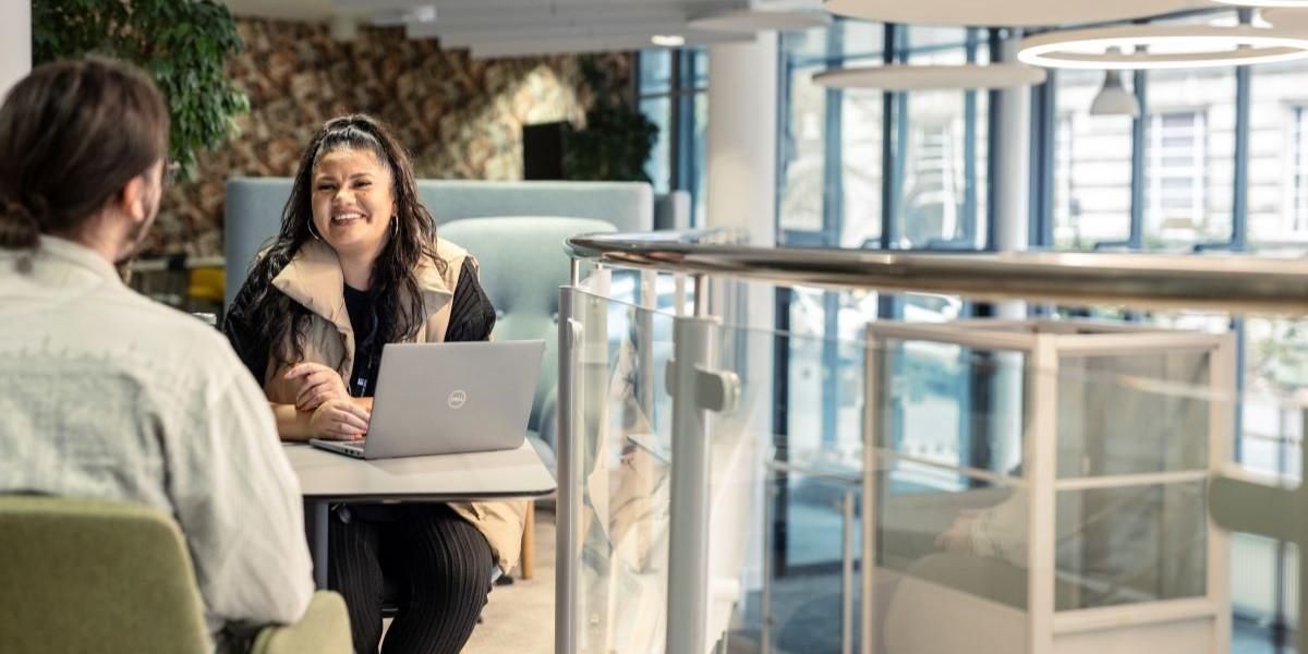 Two people sit at a table on a mezzanine floor and chat amiably. One person, who has an open laptop in front of them, laughs while the other one talks.