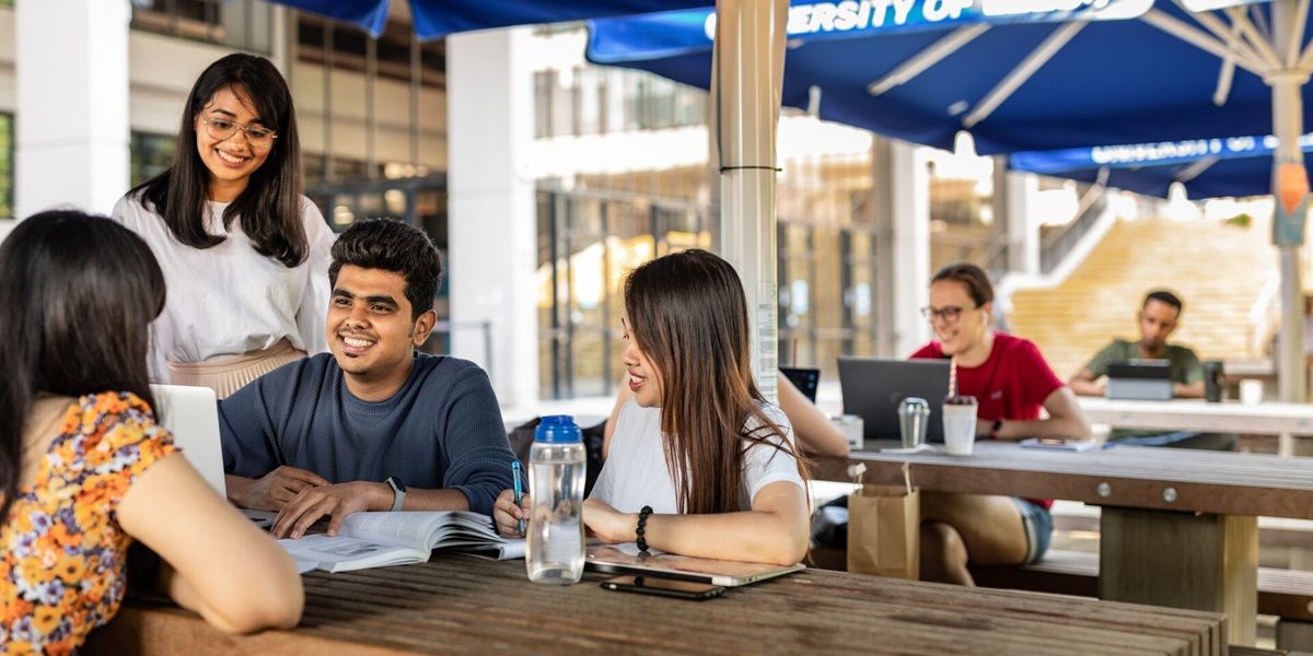 Four students chatting and using laptops at a picnic bench on a sunny day, underneath parasols.