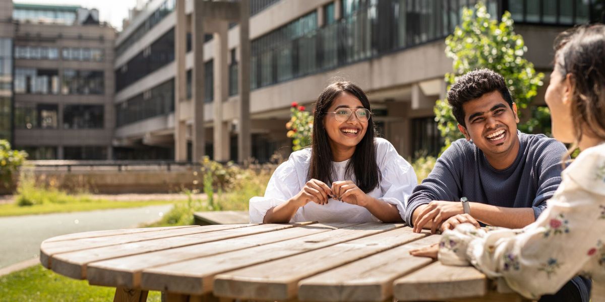 Three students sat on a picnic bench, socialising and relaxing near the Sustainable Garden on campus.