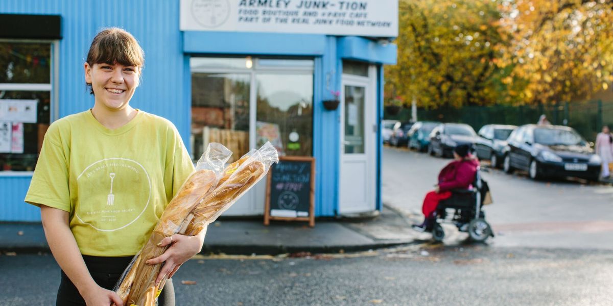 A smiling student volunteer stands holding baguettes in front of Armley Junk-tion cafe with a person in a wheelchair going up a kerb in the background.
