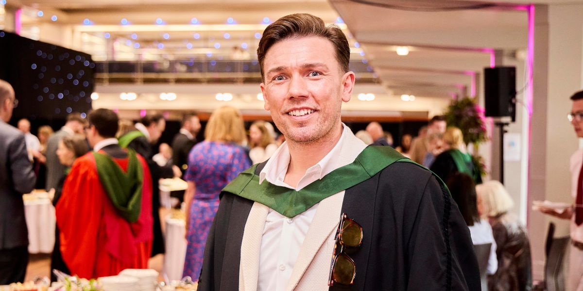 A graduate in ceremonial robes smiles at a reception with other graduates socialising among tables and a sparkling curtain.