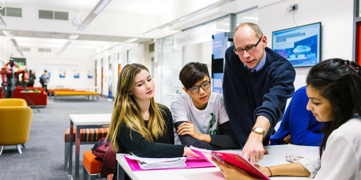 A teacher helping four students sat at a table, pointing at something in a book.