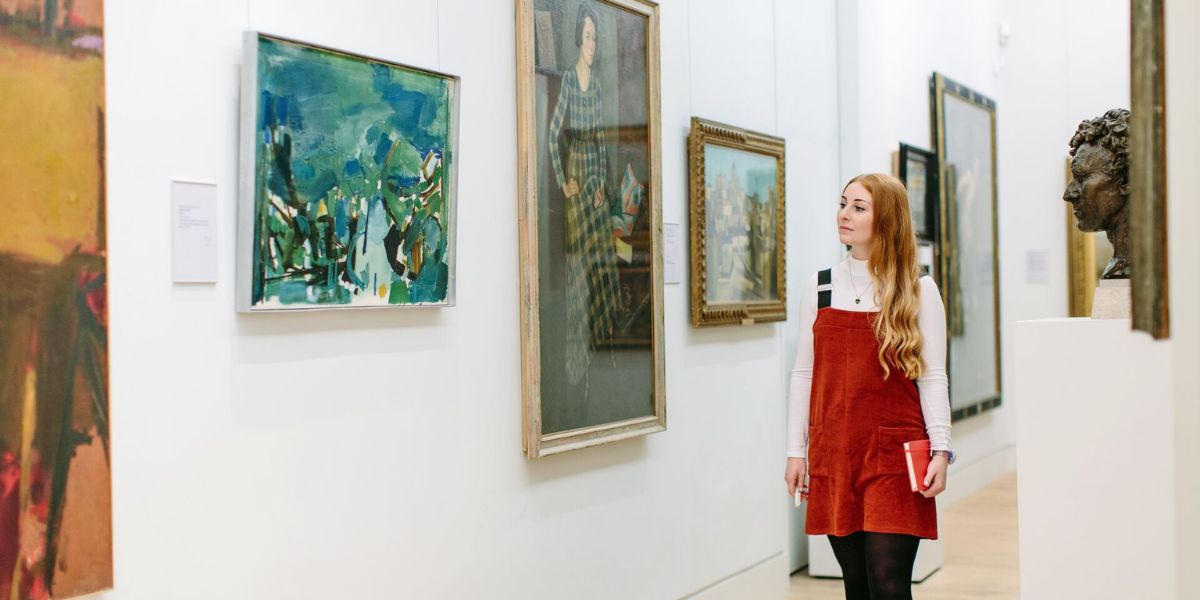A student in a red dress walking through an art gallery, thoughtfully observing paintings and sculptures on display.