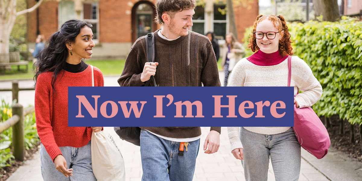 Three students walking along a path on campus, chatting and smiling. There are lots of plants and a red brick building in the background. Text says: Now I'm here.