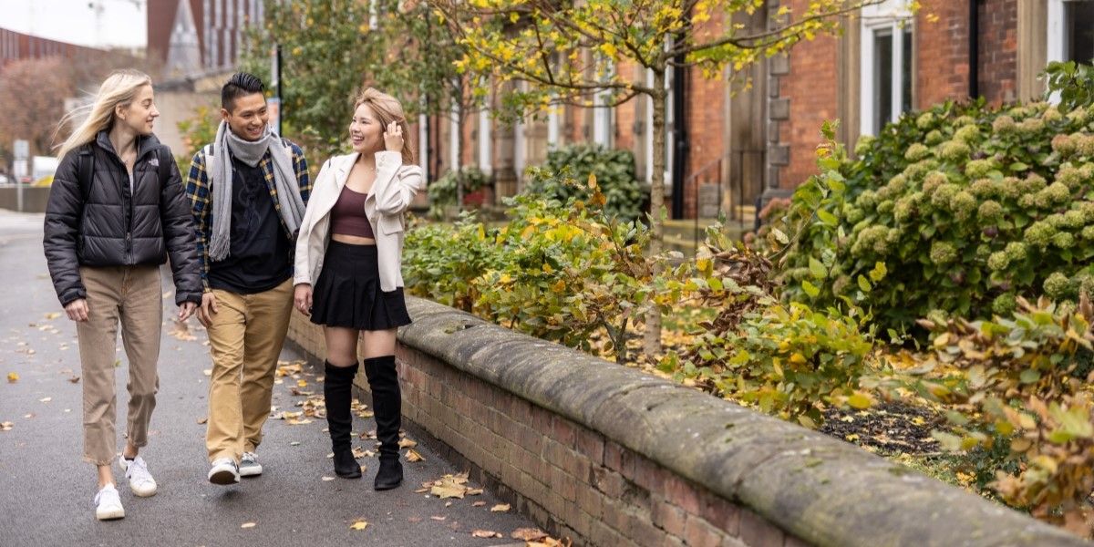 Three International Foundation Year students walk down a road on campus, talking and laughing. It is Autumn and there are golden fallen leaves on the ground.