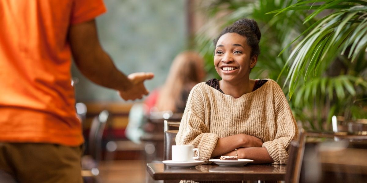 A person sat down at a table in cafe with a cup of coffee. they are chatting to someone stood up on front of them and smiling.