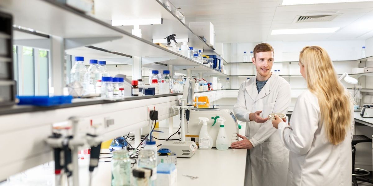Two students work inside a laboratory. Both students wear white lab coats and one passes a small petri dish to the other, who is smiling.