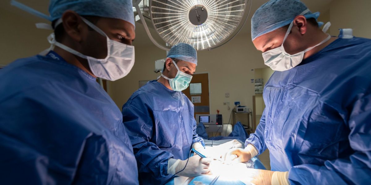 A group of surgeons perform a procedure in a sterile operating room. The surgeons are dressed in standard surgical attire, including gowns, masks, gloves, and caps and there is lighting focused on the operating table.