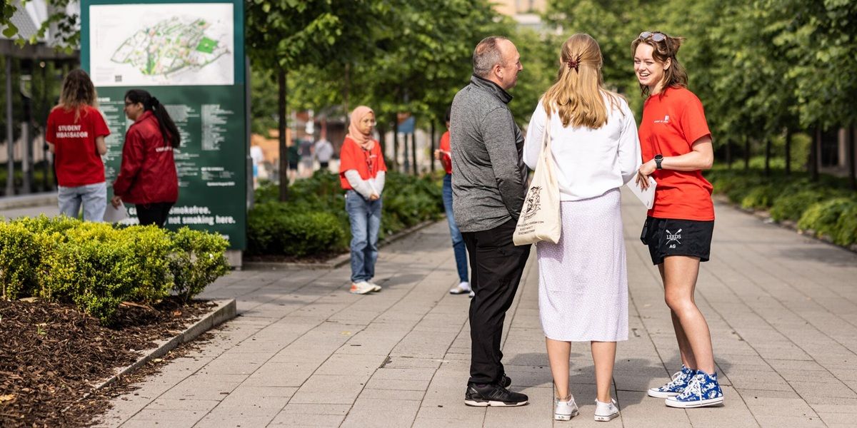 An open day ambassador in red t-shirt speaking with two open day visitors outside on campus. There are other visitors and ambassadors in the background.