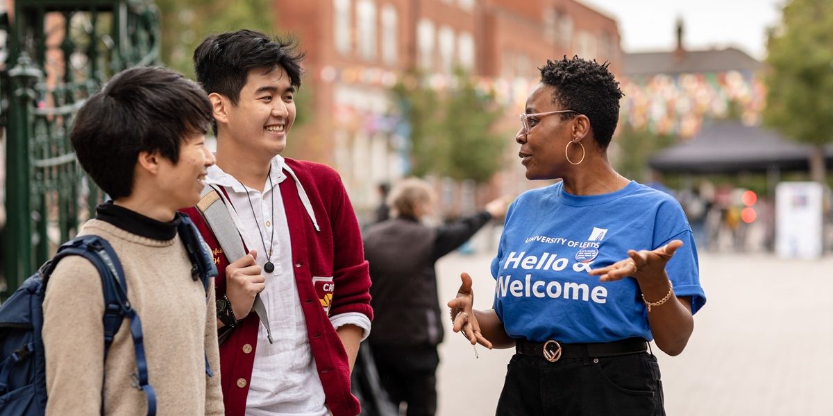 A Welcome ambassador in a blue t-shirt that says 'Hello & Welcome' on it, chatting to two smiling students on campus.