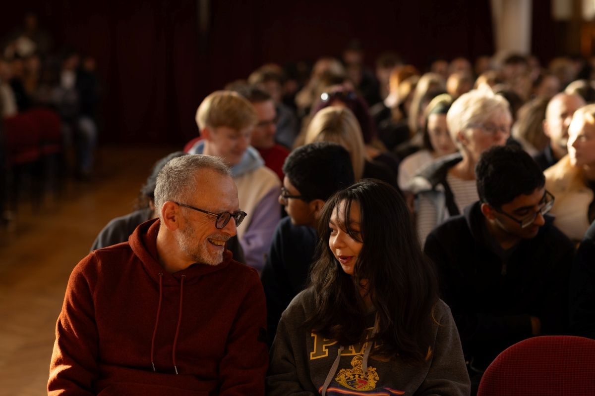 A parent and young person sit next to each other chatting. Behind them are rows of people as they wait for a talk in the Great Hall.