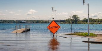 Rising sea water partially submerges a footbridge and has flooded a nearby road. There is an orange caution sign in the foreground