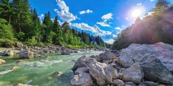 a river in full flow in the Alps, cascading over rocks in the foreground. In the background are trees and bright blue sky