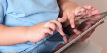 Close-up view of a small child wearing a light blue shirt, using both hands to touch and tap on a tablet device with a reflective screen