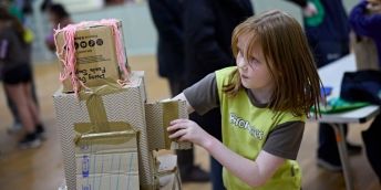 A child in Brownie uniform builds a robot using cardboard boxes, brown tape and pink strands of wool