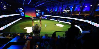 An indoor arena with a seating around the outside, a green pitch in the middle and a large screen at the end. Players are standing on the green pitch and an individual stands to clap in the stands.