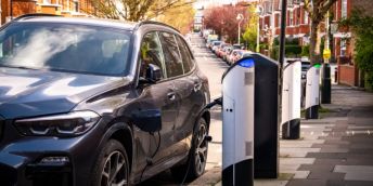 Electric car charges at a pavement charging point on a sunny suburban street. The car is in the foreground of the image, with a row of red brick houses and parked cars behind it.