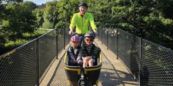 Two children wearing helmets sit in the front cargo area of a black electric cargo bike while an adult in a high-visibility jacket cycles behind them across a narrow metal bridge, surrounded by dense green trees in bright sunlight.