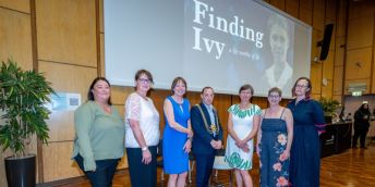 The event panellists, host and the University Vice-Chancellor stand before a screen showing the face of Ivy Angerer