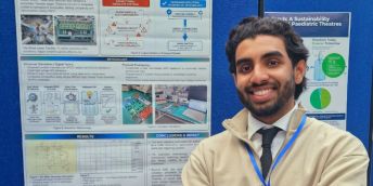 Farhan stands, smiling at the camera, in front of a blue board on which are pinned his research documentation