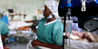 An image of a hospital ward with an IV drip in the foreground and a healthcare worker in a mask, cap and scrub standing beside a patient's bed in the background.