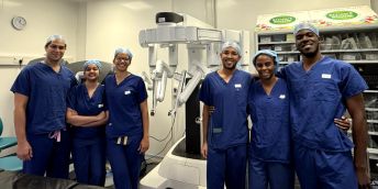 Four students with a researcher and consultant, standing in front of surgical robotics equipment in a hospital.