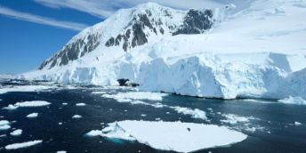 Snowy Antarctic landscape featuring a glacier, plus ice floating on the ocean, with a backdrop of blue sky.
