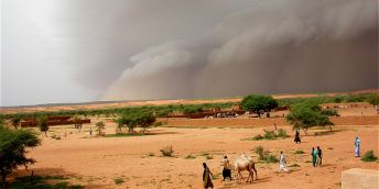 Massive storm clouds gather over a desert landscape in Mali. There are people with camels in the foreground.