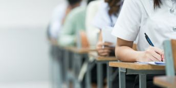 Students seated in rows at desks, writing on papers during an exam, with one student’s hand and pen in focus in the foreground.