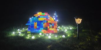 A tiny house made of colourful blocks is surrounded by fairy lights, with a miniature street lamp, on a grassy floor.