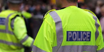 Two police officers with their back to the camera, they are wearing green hi-vis jackets with a blue and grey police badge on the back.