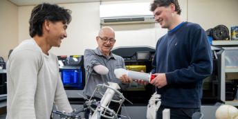 Three smiling people in a workshop examine a white prosthetic limb component beside 3D printers and a mechanical assembly on a workbench. The person on the right is holding out the prosthesis for a person with an  upper limb amputation to try on.